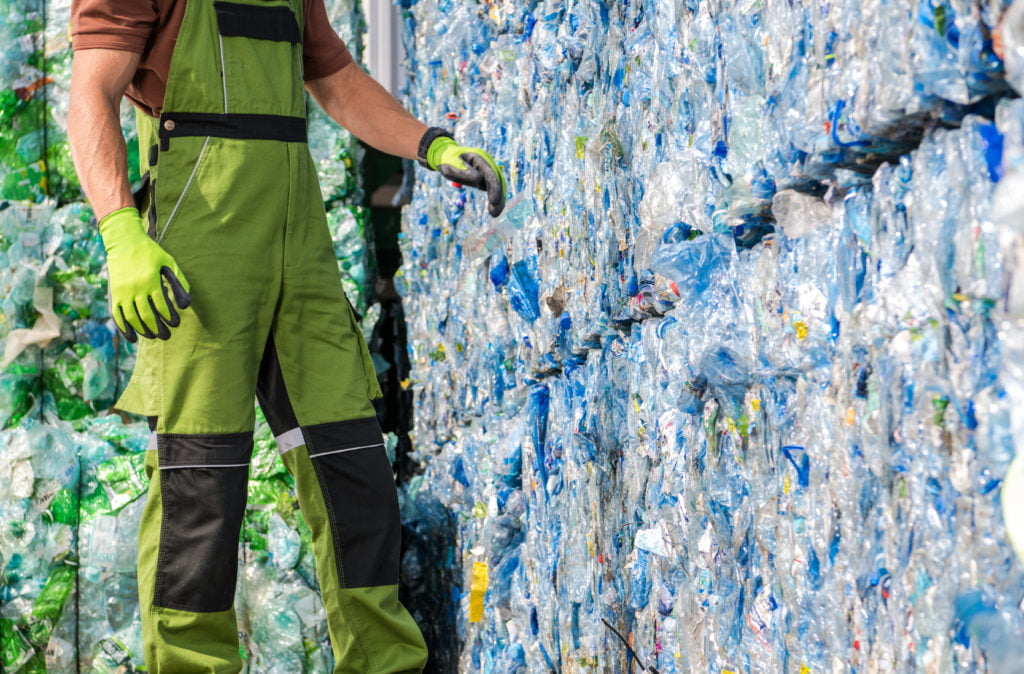 A worker in overalls is standing next to a pile of plastic bottles.