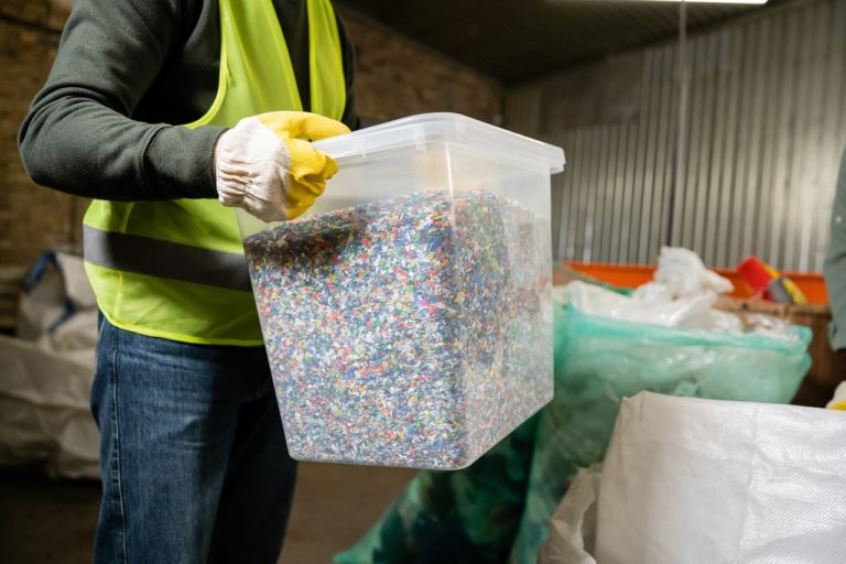 A person in a safety vest holds a clear plastic bin filled with multicolored plastic pellets in an industrial setting.